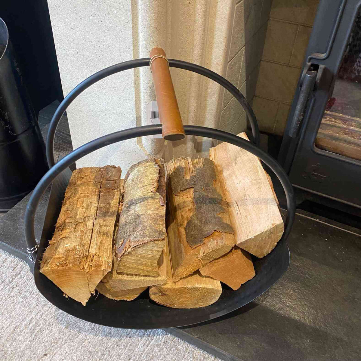 Round black iron fireside log basket with leather handles storing firewood near brick fireplace