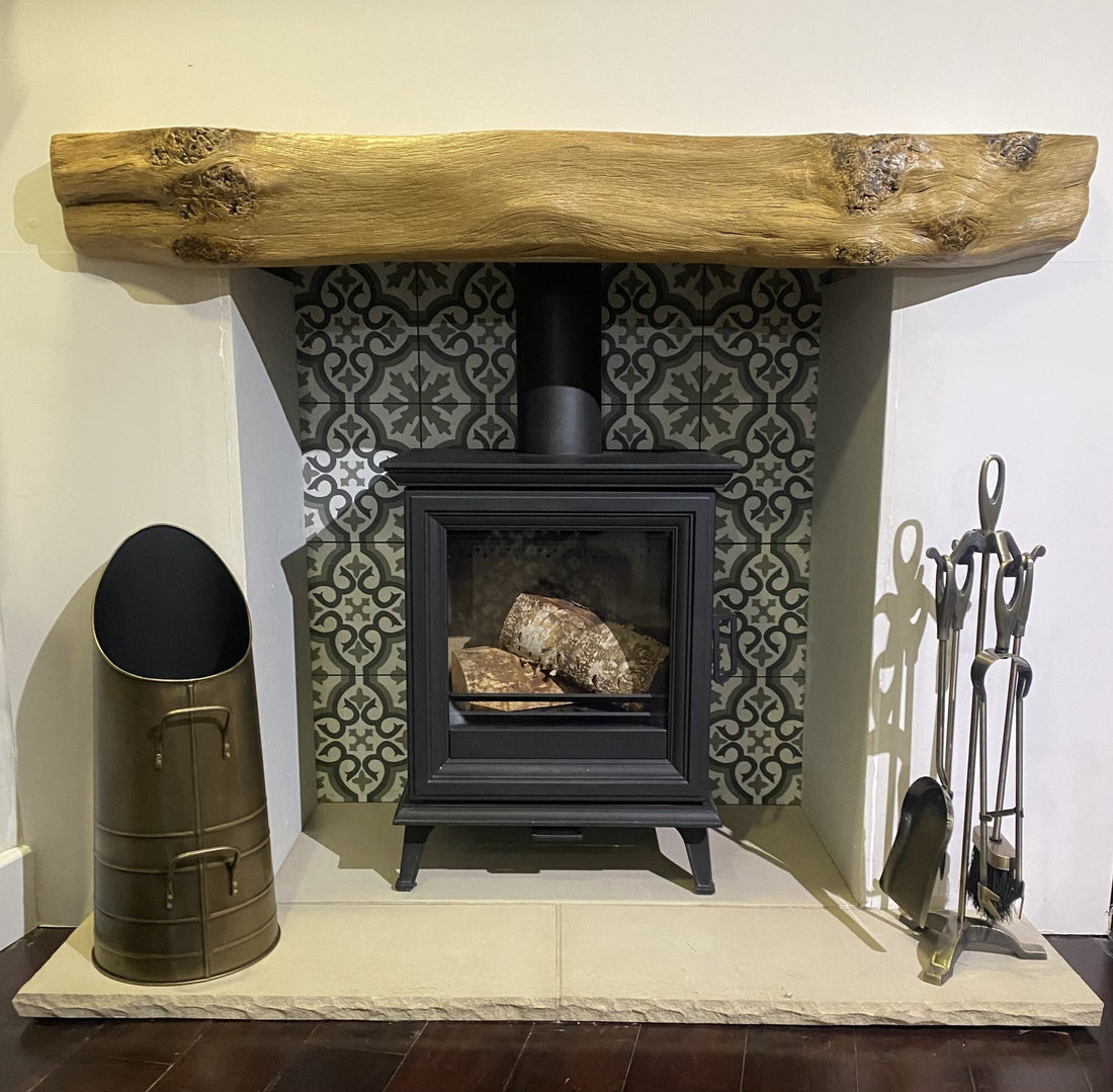 A rustic wooden fireplace mantel with natural knots and grain, mounted above a black wood-burning stove with firewood inside, set against decorative patterned tiles in a cozy home interior.