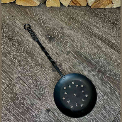 Black perforated wrought iron nut roasting pan on wood floor
