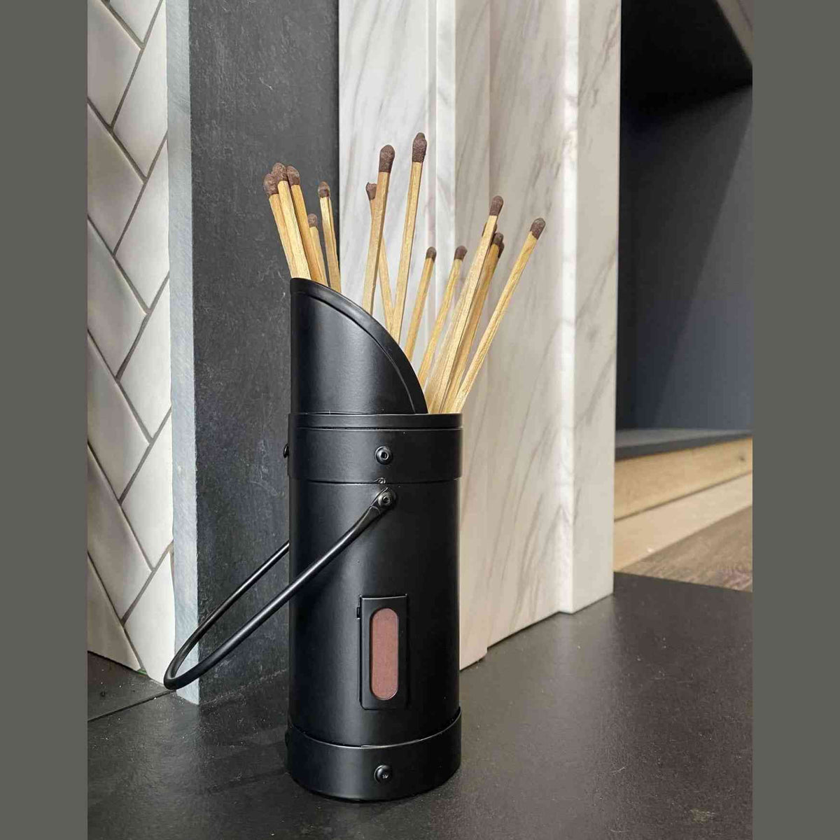 Black Calfire match canister on dark countertop beside herringbone tile fireplace