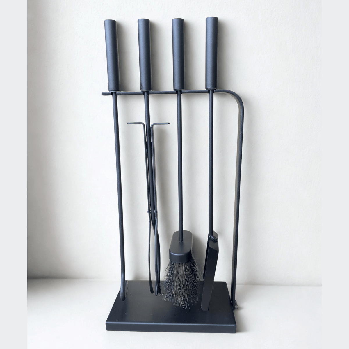 Close-up of black fireplace tools with long handles, including a poker, brush, tongs, and shovel, standing on a black hearth tool holder.
