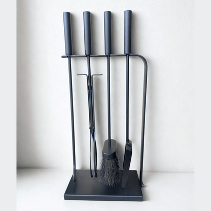 Close-up of black fireplace tools with long handles, including a poker, brush, tongs, and shovel, standing on a black hearth tool holder.