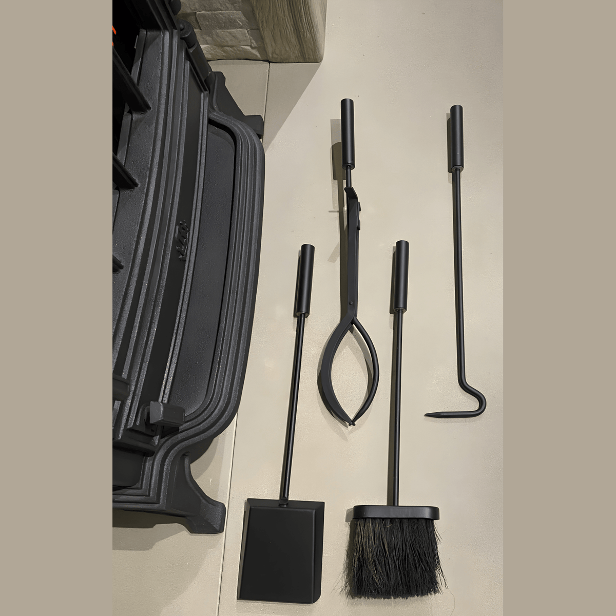 Black fireplace tool set with four long-handled tools, including a brush, tongs, poker, and shovel, on a black stand on a light-colored floor.