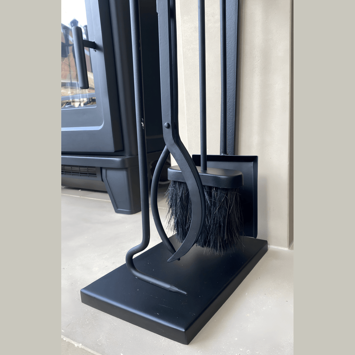 Black fireplace tool set with four tools including a brush, tongs, poker, and shovel, standing on a black base near a stove.