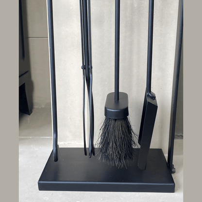 Black fireplace tool set with four tools on a black stand, including a poker, brush, tongs, and shovel, in front of a white wall.