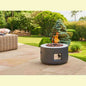 Lifestyle shot of lit grey cylindrical gas fire pit on patio with trees and sofa background