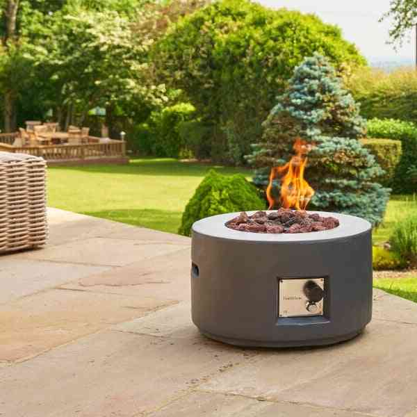 Grey outdoor gas fire pit on stone patio with flames and wicker furniture in garden setting