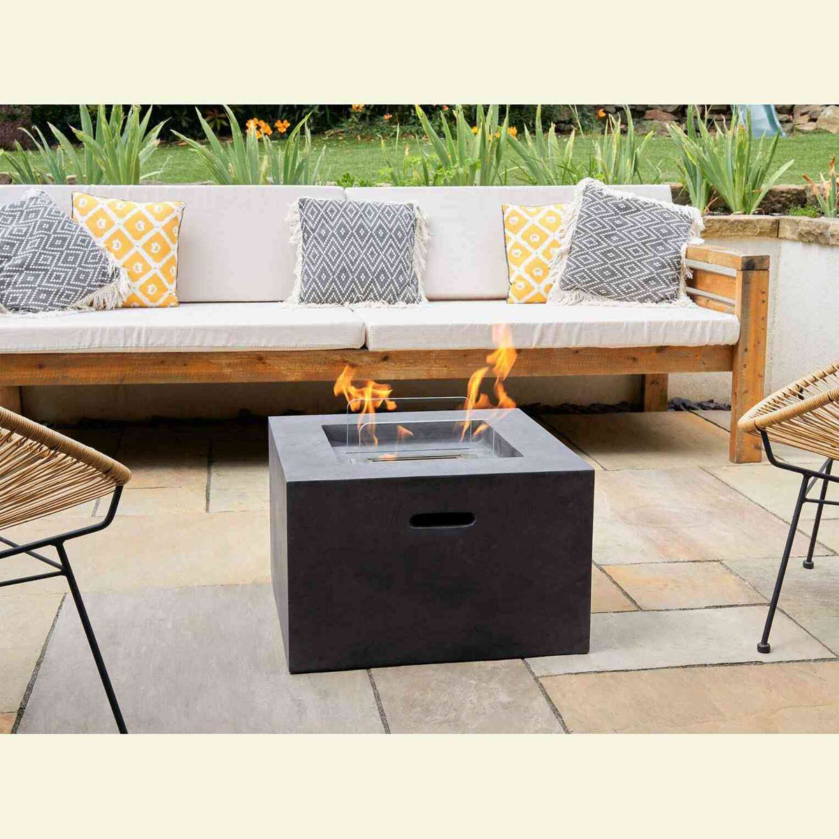 Square dark concrete grey bio ethanol fire pit lit on stone patio between wicker chairs