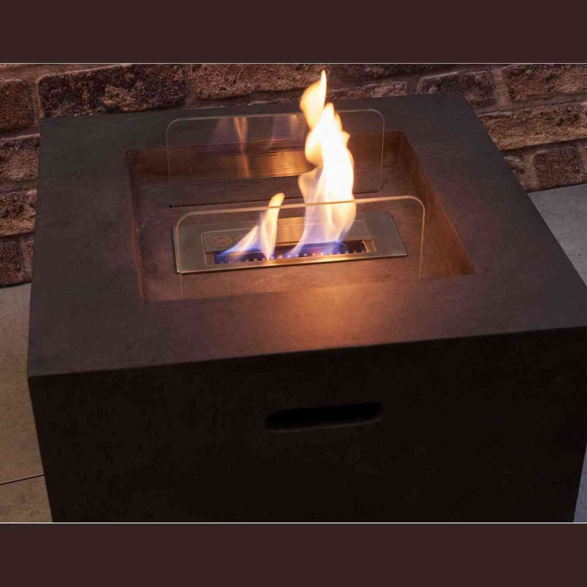 Lit dark concrete grey outdoor bio ethanol fire pit on paved patio with brick wall background