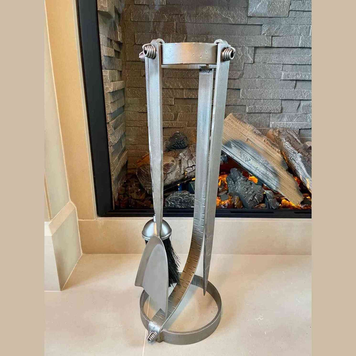 Forged steel fireside companion set on curved open-frame stand against stone-tiled wall