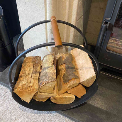Round black iron fireside log basket with leather handles storing firewood near brick fireplace