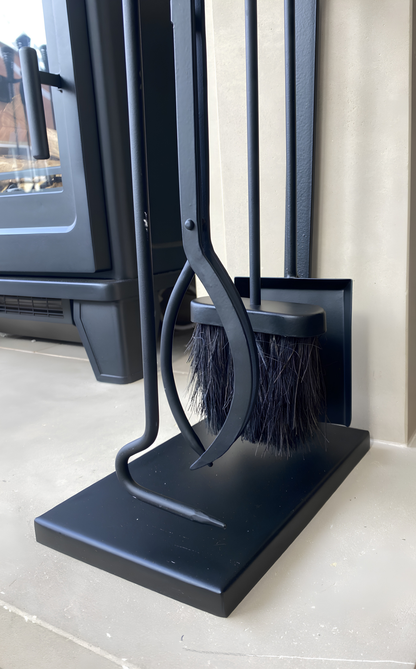 Angled side view of the Calfire Burlton 27149 black fireplace companion set positioned neatly on a floor hearth next to a contemporary log burning stove.