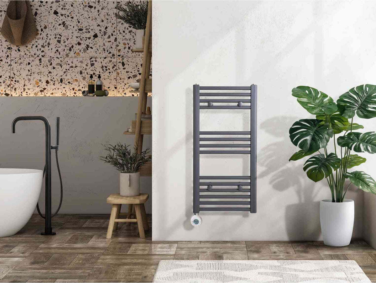 Grey electric towel radiator with digital control on a white bathroom wall, next to a large green plant in a white pot