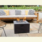Field and Flame Cassia Square Bio Ethanol Fire Pit in dark concrete grey positioned on a stone patio in front of an outdoor sofa with an open flame.