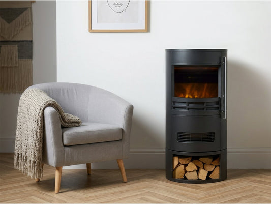 Sureflame Dunston freestanding cylindrical electric fireplace with integrated log store placed next to a grey armchair in a well-lit home interior.
